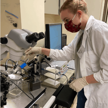 Student Kayla Gaudet working in a laboratory with a large microscope and gas chromatography set up.