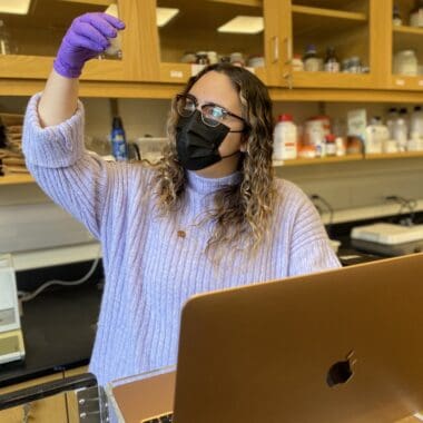 A woman in a purple sweater and purple gloves looks at a soil sample in a small white dish. She is working in a science lab.