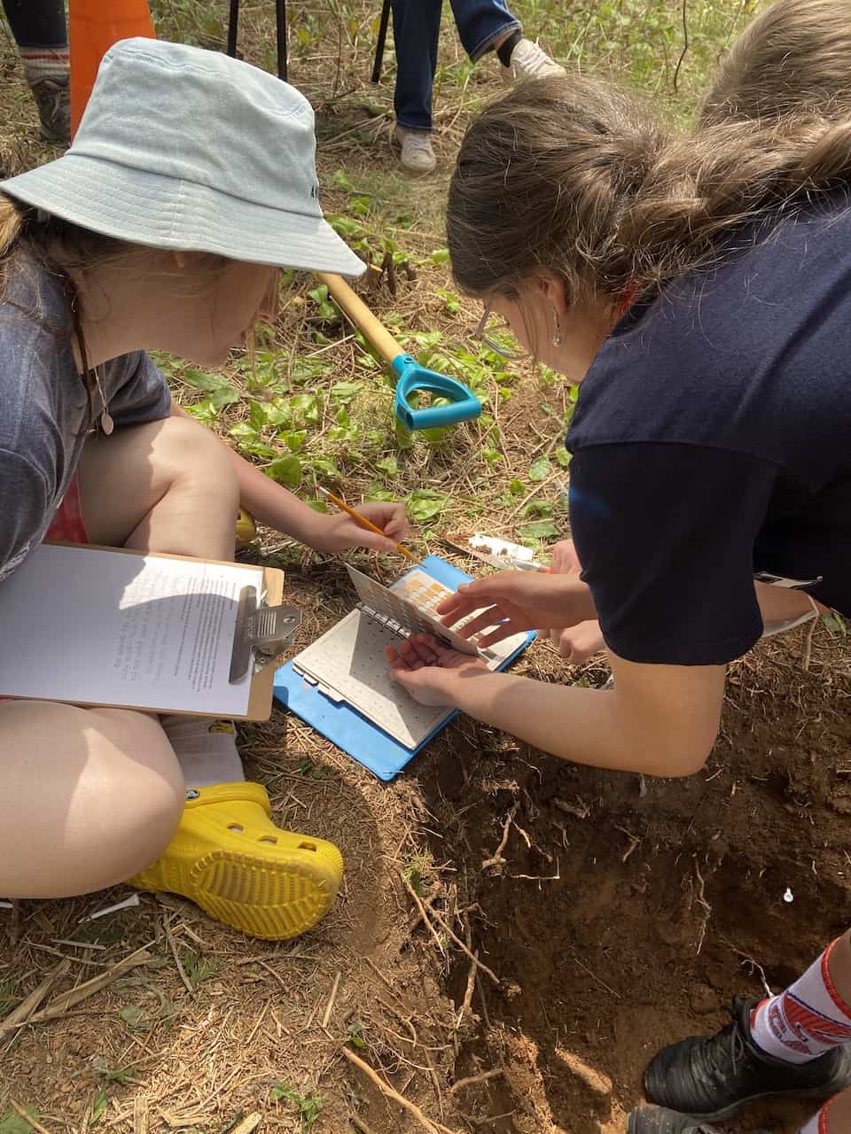 Two girls in a soil pit in the woods. They are holding a soil sample next to a book of color samples.
