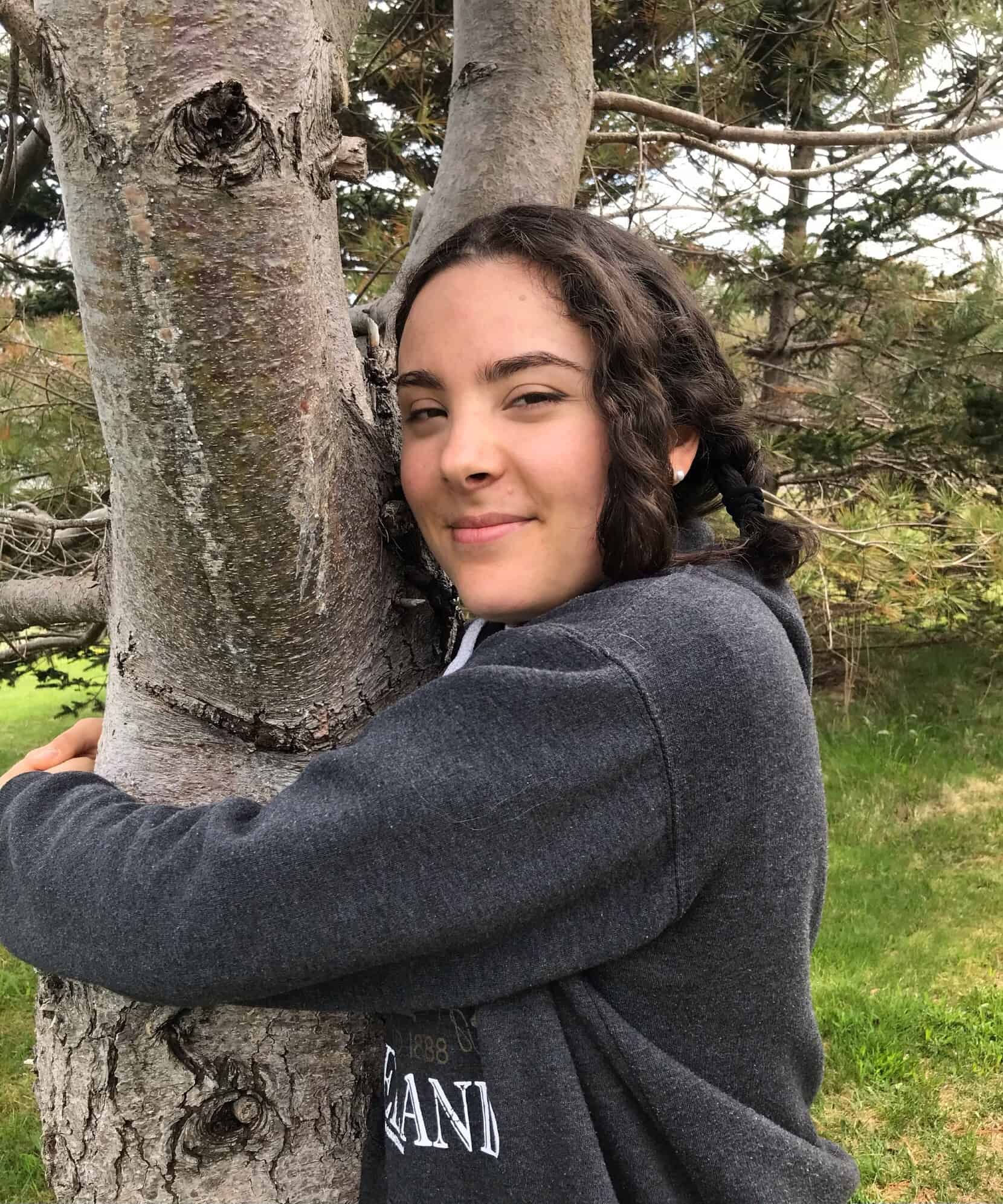 Isabelle, a young woman with dark shoulder length hair and brown eyes hugs a tree, smiling